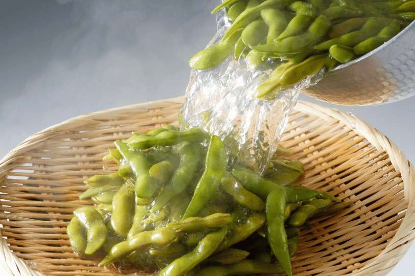 Freshly boiled edamame pods being poured from a pot into a bamboo colander to drain hot water before shocking.