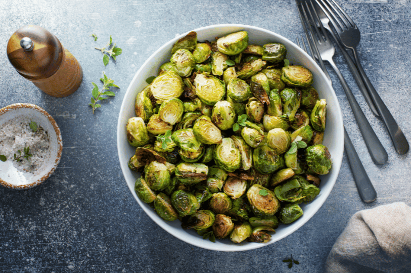 A white bowl filled with oven-roasted Brussels sprout halves, garnished with fresh thyme and served alongside a salt cellar and pepper grinder.