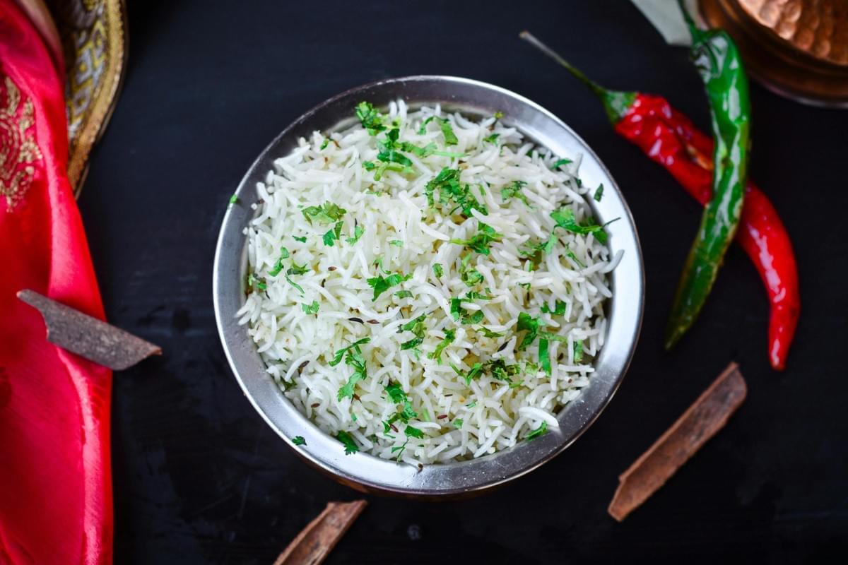 Captured from directly above, a bowl of long-grain white rice is speckled with cumin seeds and garnished with fresh chopped herbs. The presentation is framed by cinnamon sticks, red and green chilies, and peppercorns scattered on a dark surface next to a bright red cloth.