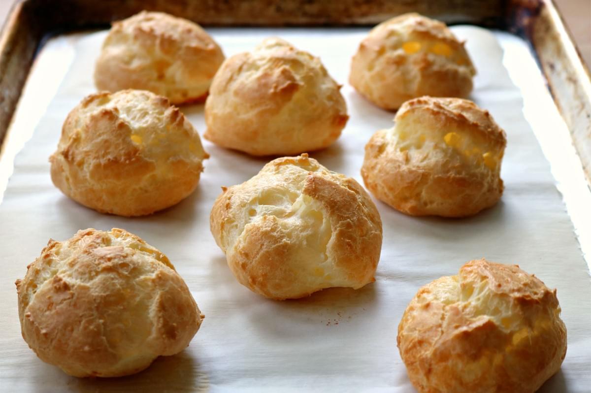 Seven large, puffy, golden-brown choux pastry shells arranged on a baking sheet lined with white parchment paper, ready to be filled.