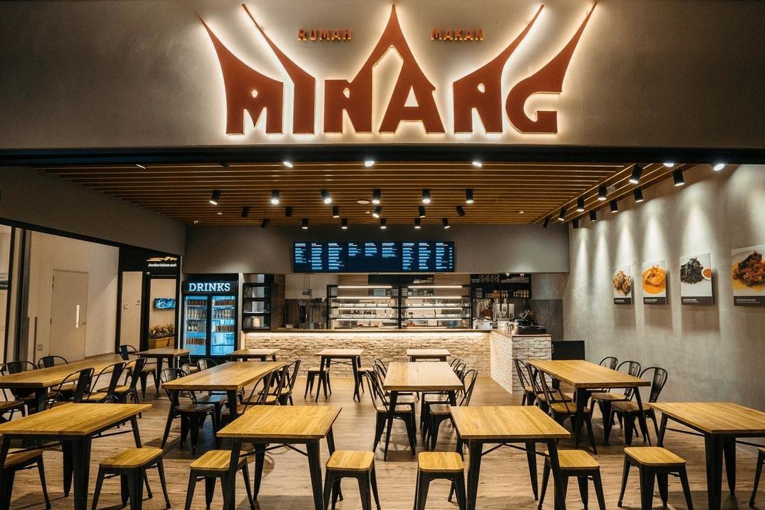 This image shows the interior of "Rumah Makan Minang," a modern Indonesian restaurant featuring warm wood tables, industrial-style seating, and a prominent illuminated logo. The space is well-lit with sleek track lighting and displays several framed food photos on a minimalist grey wall.