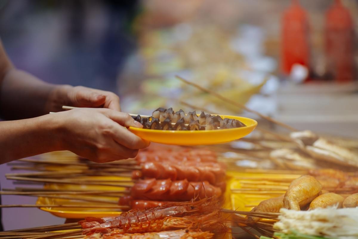 A close-up shot captures a person's hands holding a yellow plate with skewers of sliced meat or seafood at a bustling street food market. In the foreground and background, various other skewered items like sausages, prawns, and chicken wings are stacked on trays, creating a warm, vibrant atmosphere.