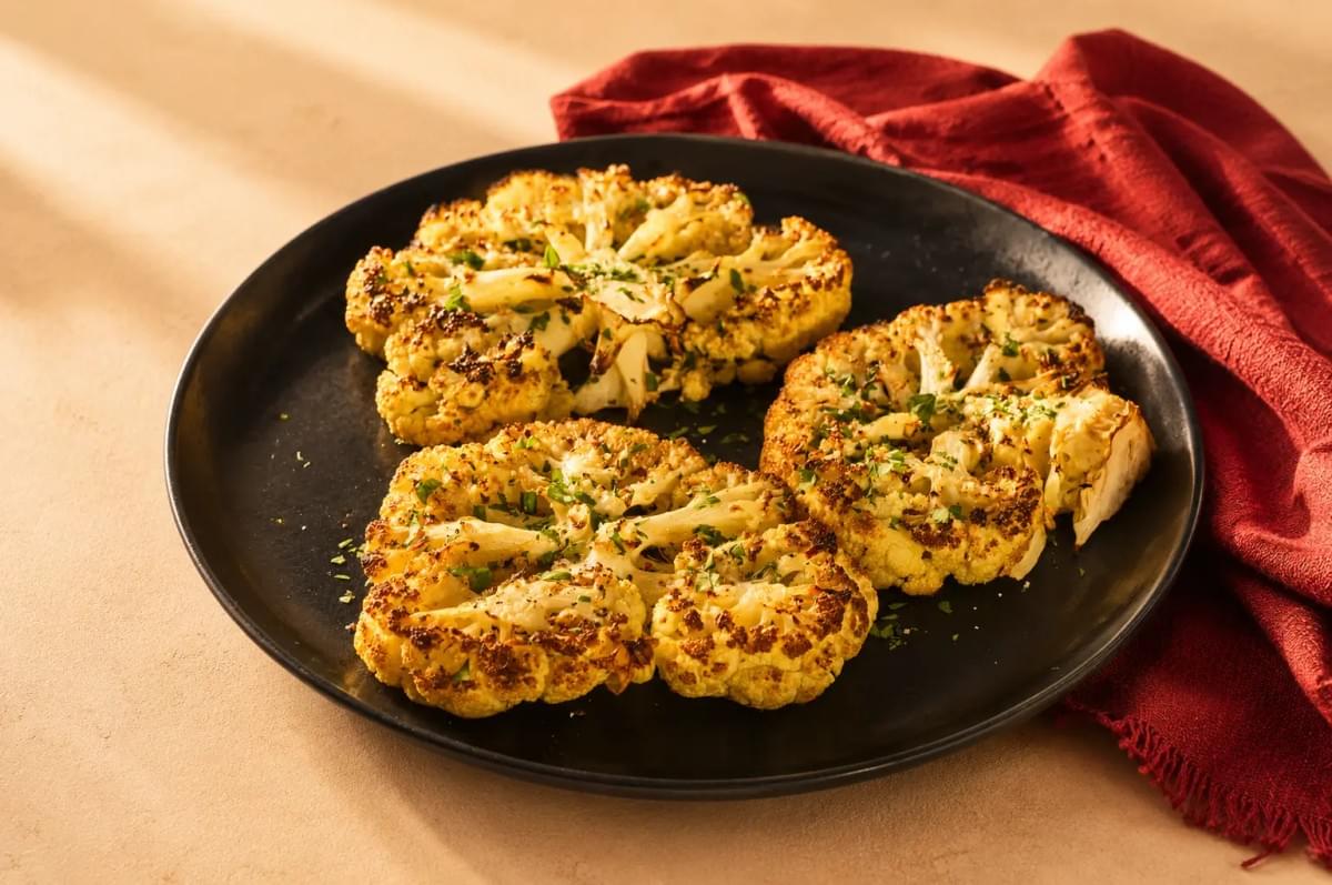 Three roasted cauliflower steaks garnished with chopped herbs, served on a black plate next to a red cloth napkin.