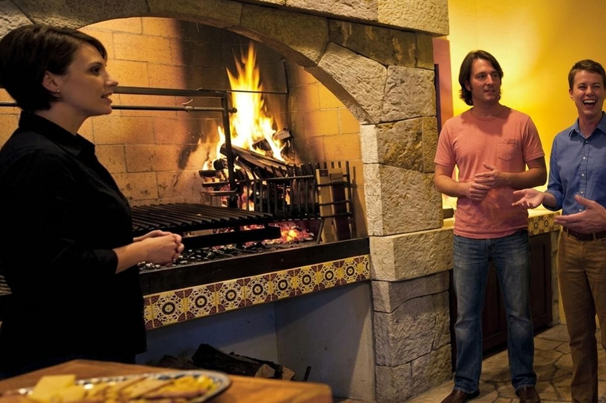 People gathering and conversing around stone asado grill with fire burning while woman tends to the grilling