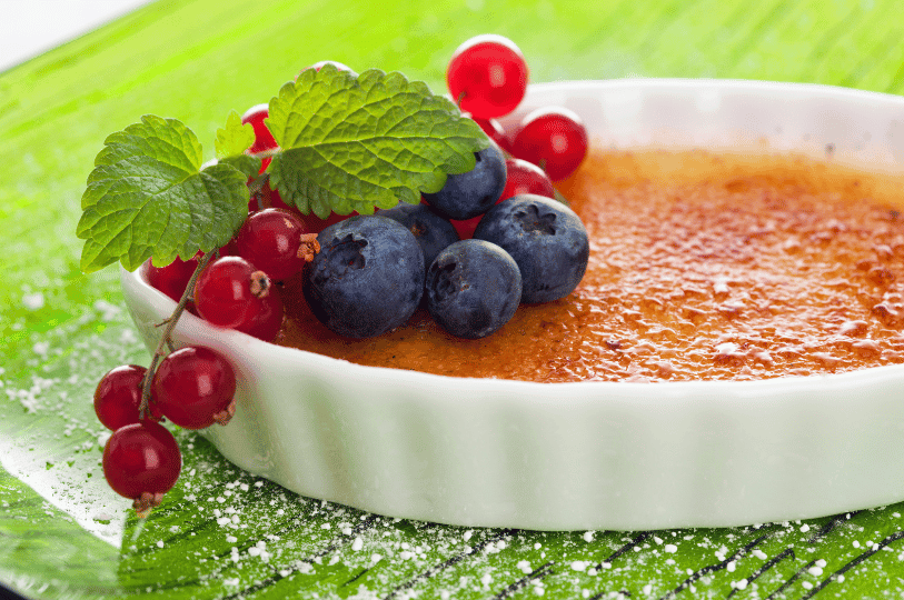 A shallow dish of crème brûlée garnished with fresh blueberries, red currants, and mint leaves, resting on a vibrant green surface sprinkled with powdered sugar.