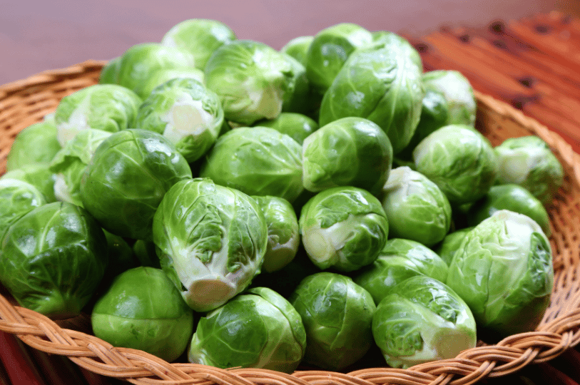 A rustic wicker basket overflowing with a large mound of whole, vibrant green raw Brussels sprouts.