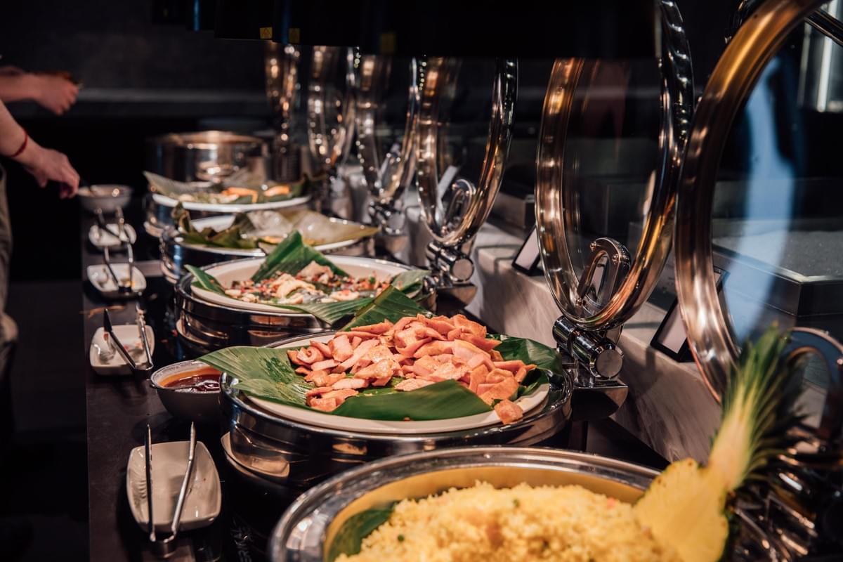 A buffet line displays a row of stainless-steel chafing dishes lined with banana leaves and filled with dishes like fried rice and cooked meat. Polished glass lids stand open under warm lighting as a guest serves food in the background.