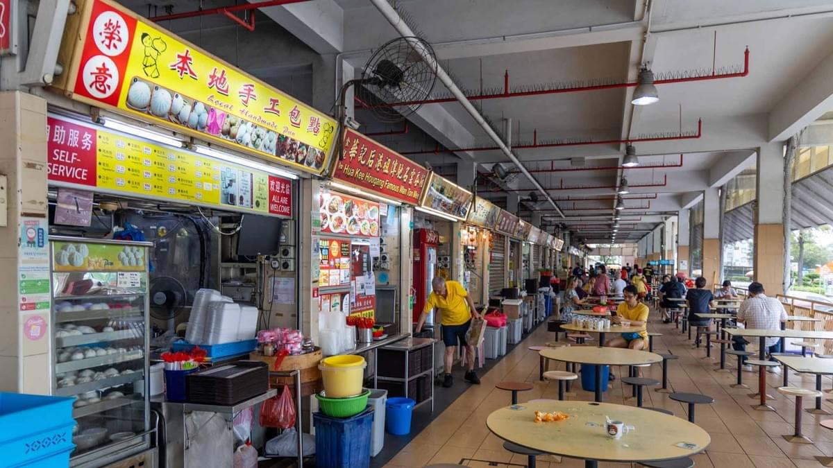This image shows a row of food stalls at the Old Airport Road Food Centre, featuring a prominent stall selling local handmade buns. Patrons are seated at round tables in the open-air dining area, with several other stalls and a vendor in a yellow shirt visible in the background.