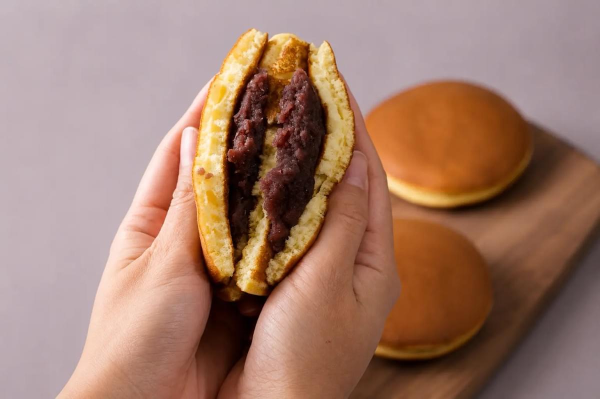 A close-up shot of a hand holding a dorayaki pancake sandwich with a large bite taken out, showcasing the thick sweet red bean paste inside the fluffy, golden interior. In the background, more whole and halved dorayaki sit on a light pink plate against a neutral tabletop.