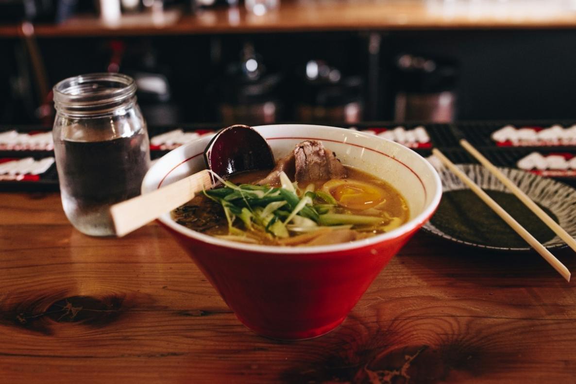 A steaming bowl of ramen with a soft-boiled egg, sliced pork, and green onions sits on a wooden counter next to a glass of water and chopsticks. The close-up shot captures the rich broth and fresh toppings in a vibrant red bowl, set against a softly blurred bar background.
