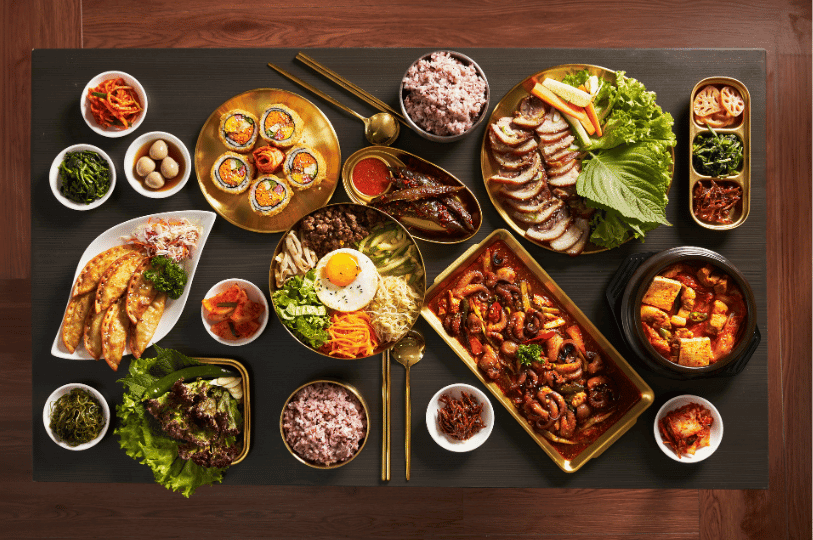 An expansive, overhead view of a lavish Korean feast spread across a dark table, featuring a central bowl of bibimbap, spicy stir-fried octopus, braised pork trotters, and fried dumplings, all anchored by numerous small bowls of vibrant banchan scattered throughout the setting.