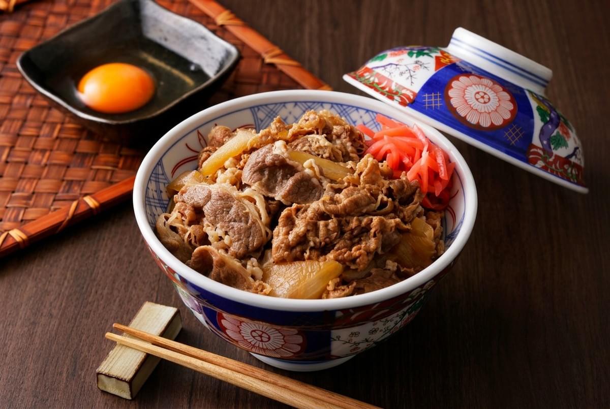 Japanese gyudon beef rice bowl served with pickled ginger, a raw egg yolk, and chopsticks.