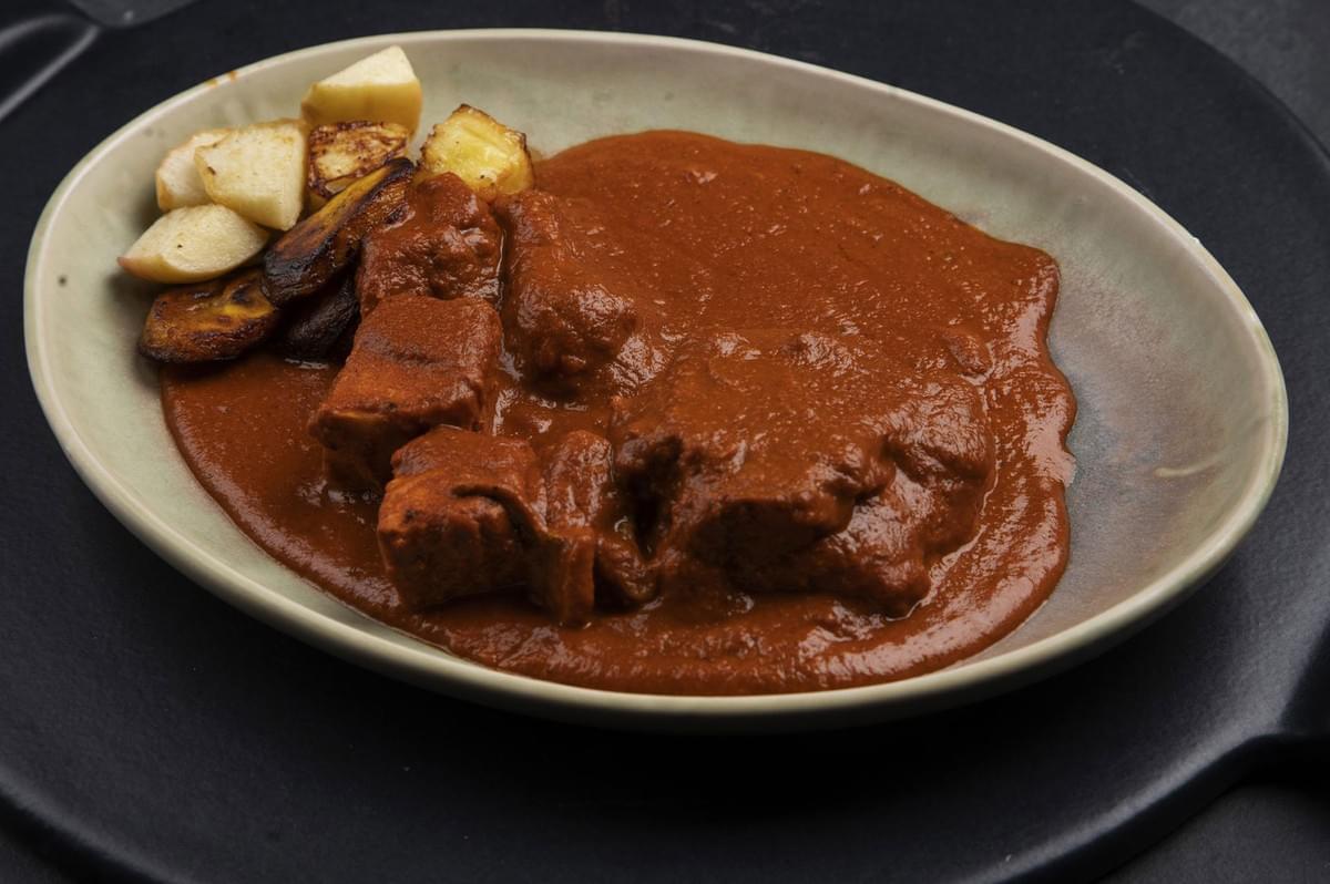 A portion of cubed pork or meat covered in a thick, rich red sauce (possibly Mole Colorado or Adobo) served with roasted potato and plantain chunks on an oval, rustic plate.