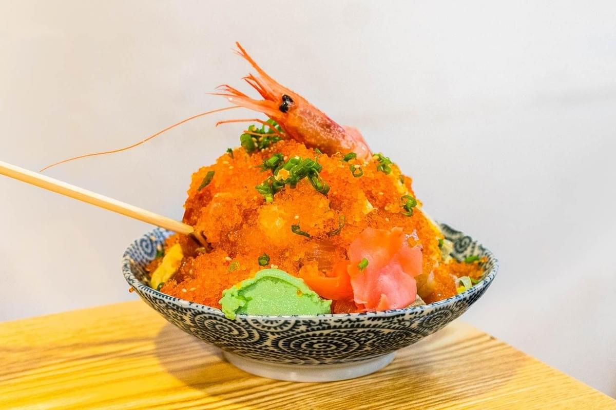 A patterned ceramic bowl sits on a wooden counter, filled with a high mound of chirashizushi topped with bright orange tobiko, green scallions, wasabi, and pickled ginger. A whole sweet shrimp rests at the top of the seafood pile, with wooden chopsticks leaning against the side.