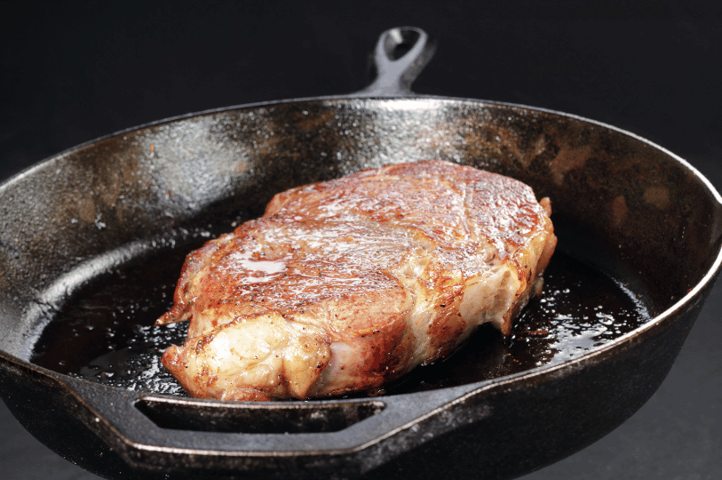 A thick ribeye steak searing in a heavy cast-iron skillet, showing the early stages of a golden-brown Maillard reaction on the surface.
