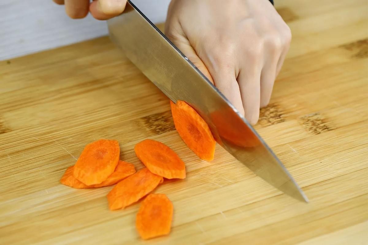 Close-up of a hand holding a large kitchen knife to cut a carrot on an angle (the bias cut) into oval-shaped slices on a light bamboo cutting board.
