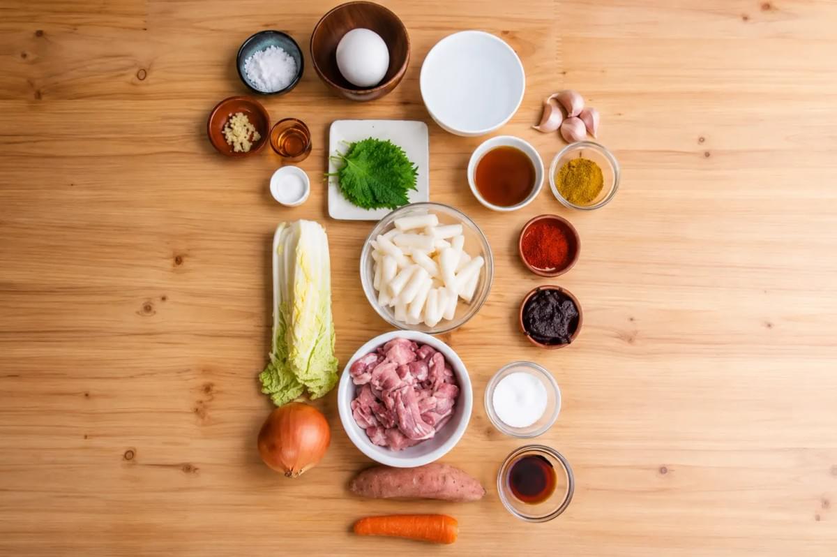 An overhead view of raw cooking ingredients neatly arranged on a wooden table, featuring chopped meat, rice cakes, napa cabbage, vegetables, and small bowls of sauces and spices.