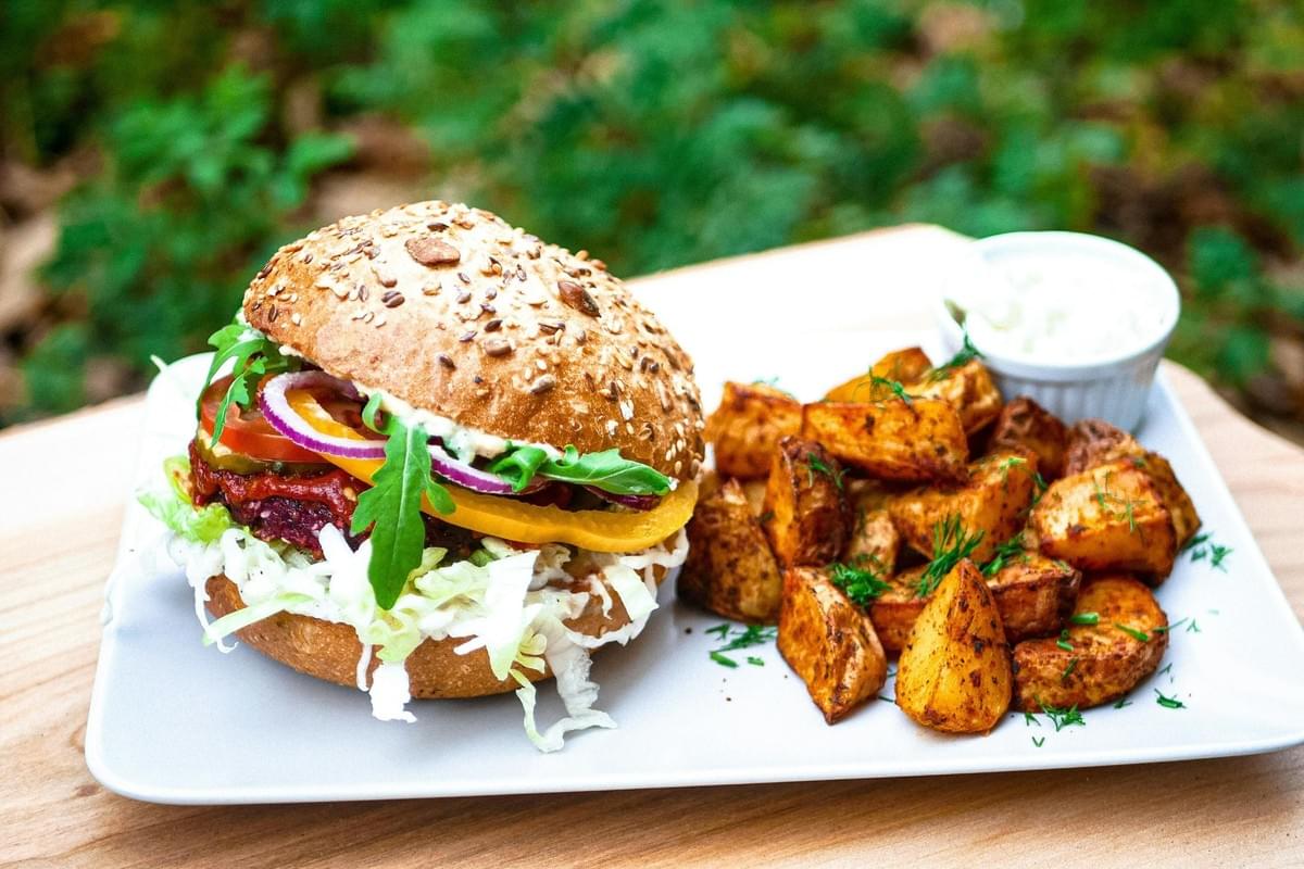 A vibrant veggie burger on a seeded bun is packed with fresh arugula, bell peppers, and red onions, served alongside a pile of seasoned potato wedges. The meal is presented on a white plate against a blurred outdoor background, accompanied by a small side of creamy dipping sauce.