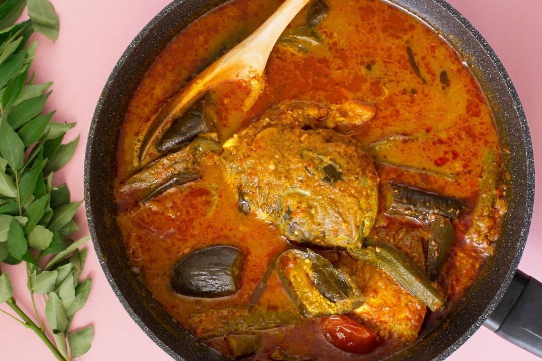 The image features a top-down view of a rich, red fish head curry simmering in a large black pan against a pink background. A wooden spoon rests inside the dish, which is packed with eggplant, okra, tomatoes, and a sprig of fresh curry leaves on the side.
