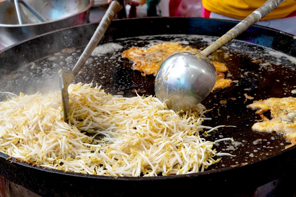 A large mound of fresh white bean sprouts is being stir-fried in a massive black wok, sizzling alongside crispy, golden-brown omelets bubbling in hot oil. A sturdy metal spatula and ladle rest directly in the pan, ready to mix the ingredients for this savory street food dish.
