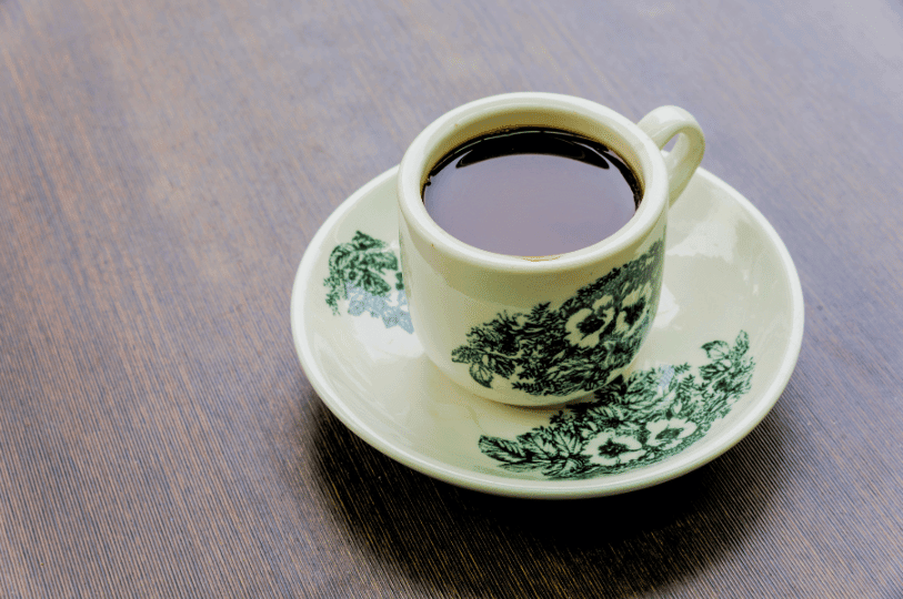 A traditional thick ceramic cup and saucer featuring green floral patterns, filled with dark black, strong Singapore-style coffee (Kopi O), sitting on a dark wooden surface.