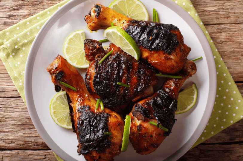 Top-down view of grilled Jamaican jerk chicken drumsticks with visible char marks, garnished with lime slices and chives, served on a white plate over a green polka-dot napkin.