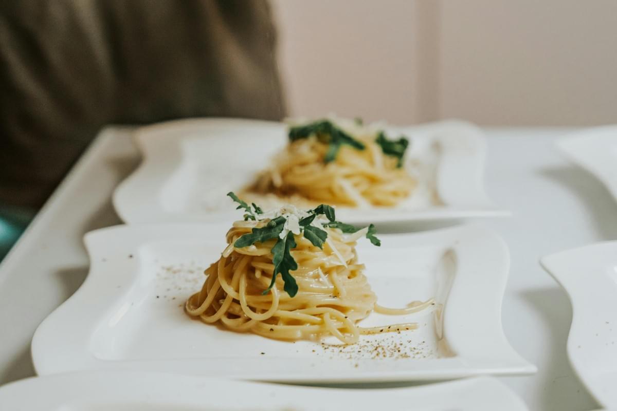 A neat mound of spaghetti coated in a creamy sauce is presented on a white, wavy-edged plate, topped with fresh green arugula and grated cheese. The plate in the foreground is dusted with black pepper, while similar servings are visible in the soft-focus background.