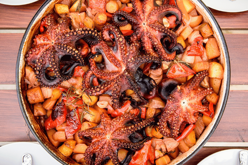 Top-down view of a vibrant seafood Peka dish containing whole roasted octopuses spread over a bed of diced potatoes, carrots, red peppers, and sprigs of rosemary.
