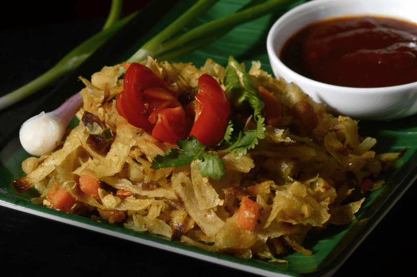 A serving of Kottu Roti on a green leaf-lined plate, topped with fresh tomato wedges and cilantro, accompanied by a small bowl of spicy red gravy.