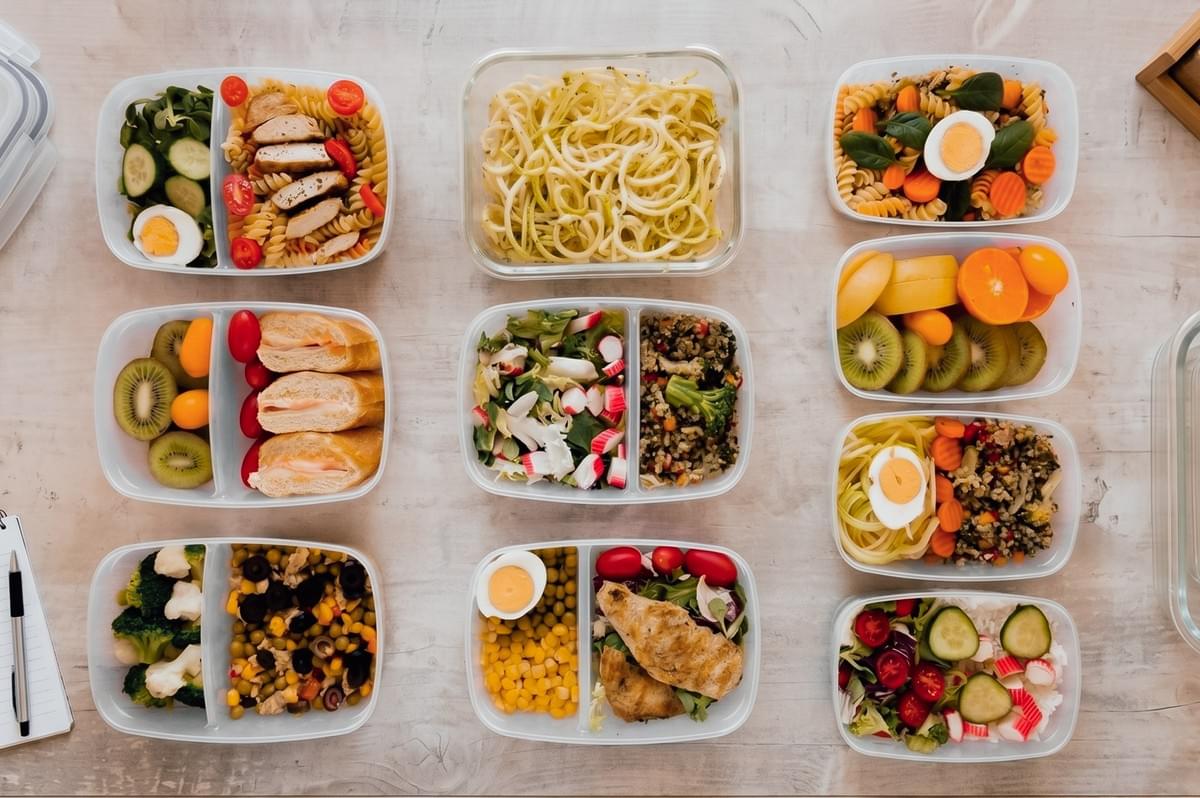 Top-down flat lay shot of multiple meal prep containers with balanced portions of pasta, vegetables, fruit, and protein, demonstrating portion control and meal planning to reduce food waste and improve food storage efficiency.