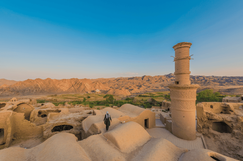 An panoramic view of an ancient Silk Road outpost featuring traditional mud-brick architecture and a tall, patterned minaret. A figure walks along the domed rooftops, overlooking a valley with sparse greenery and rugged mountains, representing the historical trade hubs where spices were exchanged.