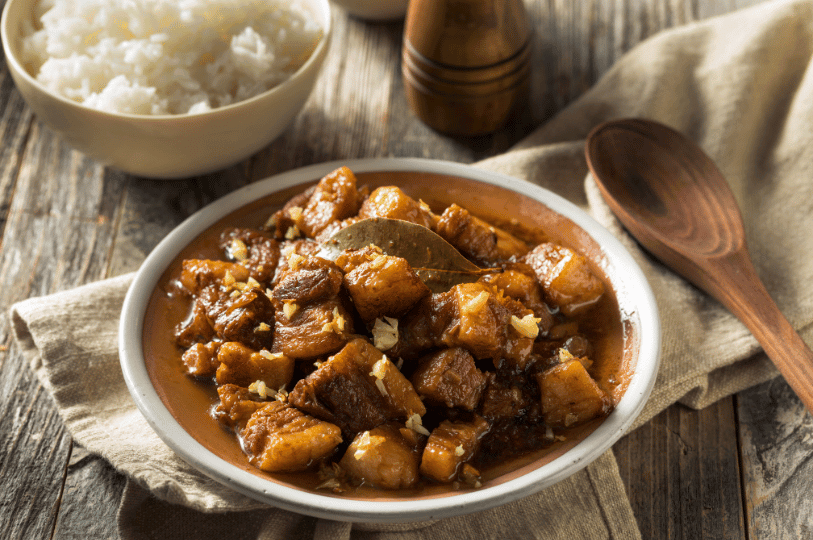 A wide rustic shot of Filipino Pork Adobo on a ceramic plate, served alongside a bowl of steamed white rice and a wooden spoon on a linen napkin.