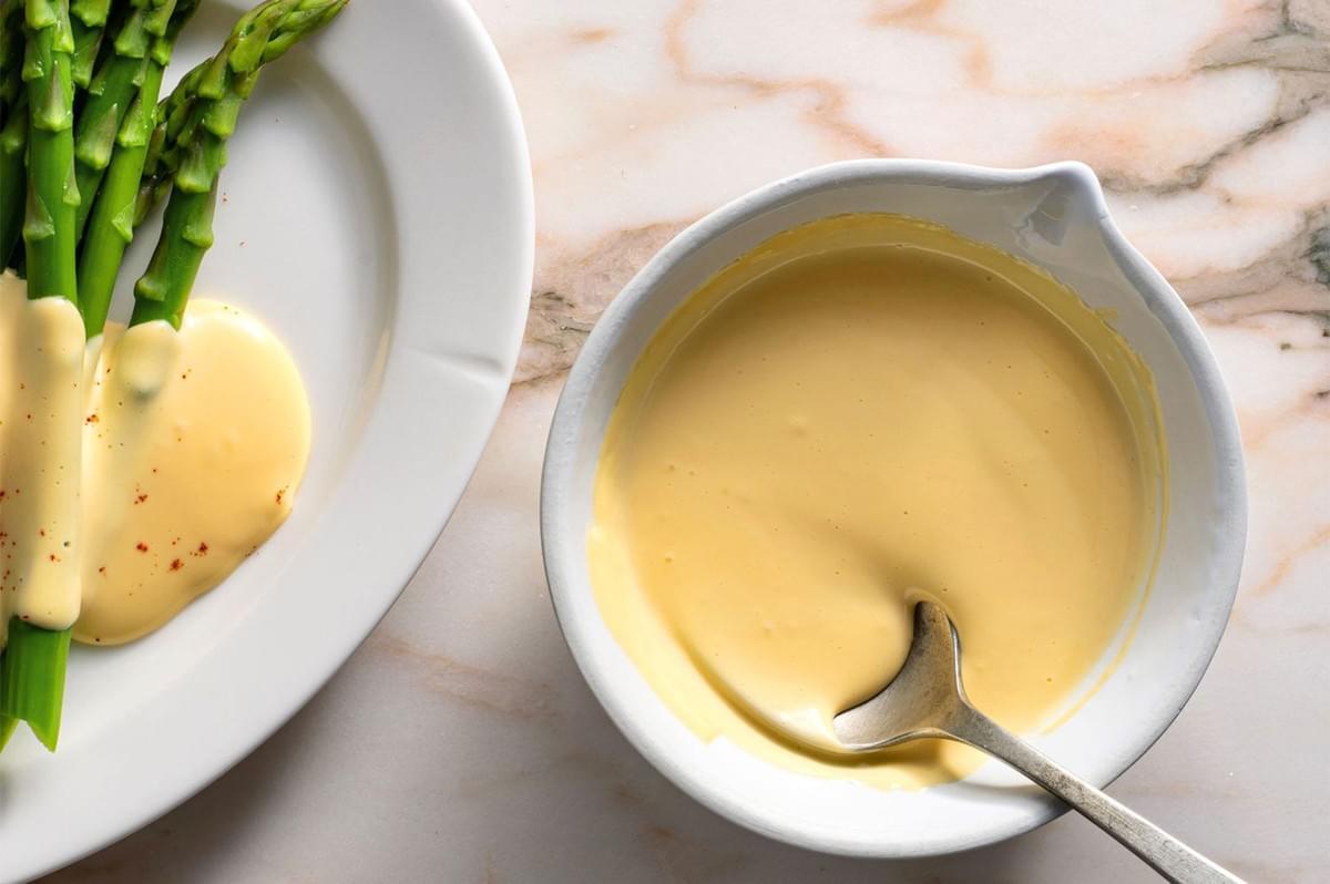 An overhead shot of blanched green asparagus spears on a white plate, partially covered with creamy yellow hollandaise sauce, with a small bowl of the remaining sauce and a spoon next to it on a marble counter.
