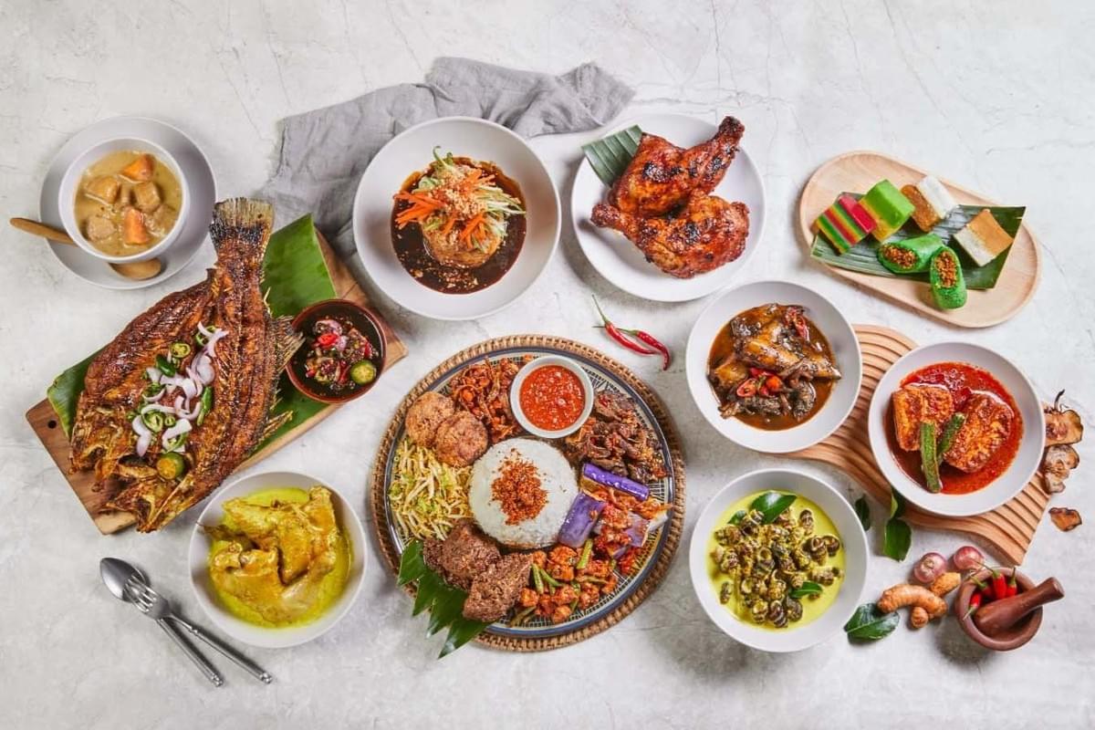 This image displays a vibrant spread of traditional Nasi Padang and Malay dishes arranged on a white marble surface, featuring a large central platter of rice surrounded by various bowls of curries and grilled meats. The assortment includes signature items like grilled fish, ayam bakar, and beef rendang, alongside colorful kuih-muih desserts and fresh aromatics like chilies and turmeric.