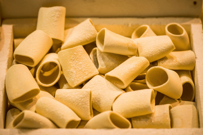 A wooden box filled with dry paccheri, a large, wide, and smooth tubular pasta shape originating from the Campania region.