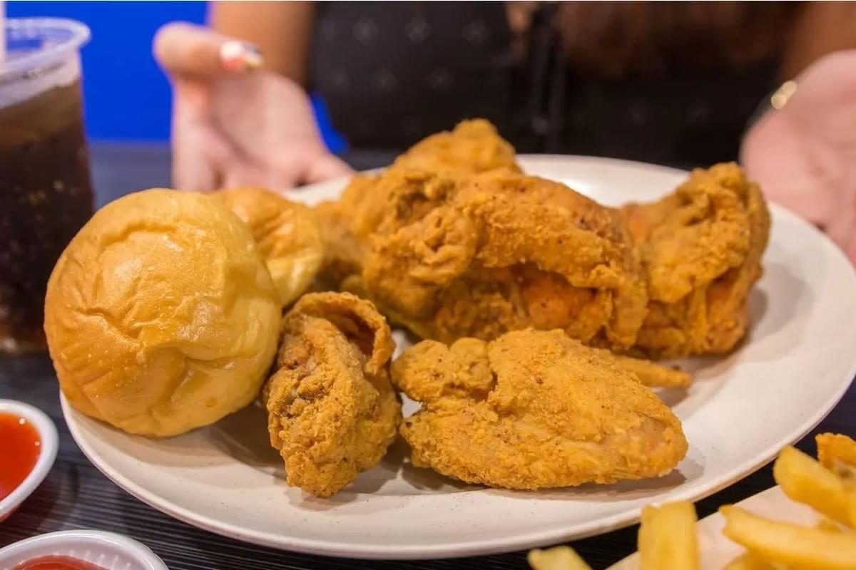 A large white plate holds several golden-brown pieces of crispy fried chicken alongside two fluffy dinner rolls. In the background, a person's hands are visible next to a cup of soda, with small dishes of dipping sauce and French fries framing the table.