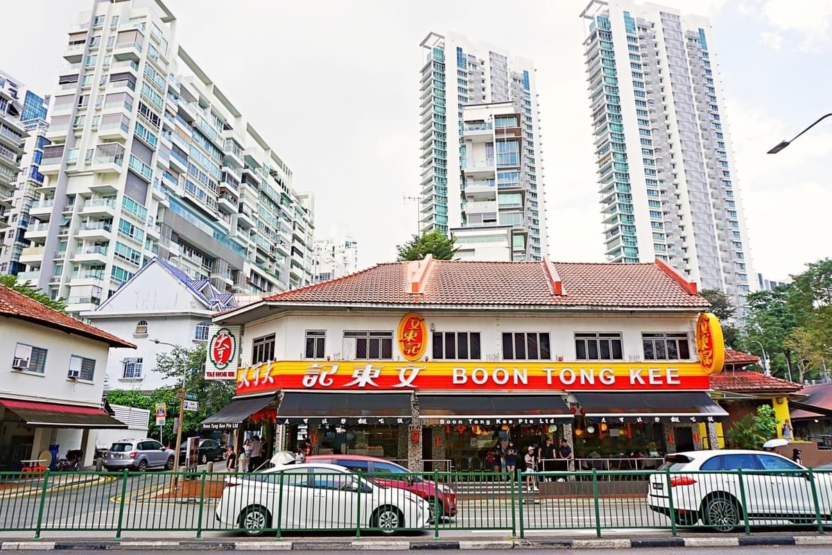 This image features a two-story Boon Tong Kee restaurant building with a distinctive red and yellow signboard, situated on a street corner against a backdrop of modern high-rise apartments. Several cars are parked or passing in front of the establishment, which is separated from the main road by a green metal fence.
