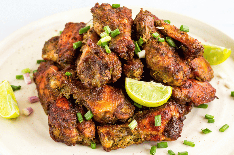 A heap of spicy, oven-roasted jerk chicken wings seasoned with aromatic herbs, topped with chopped green onions and served with fresh lime wedges on a light-colored plate.