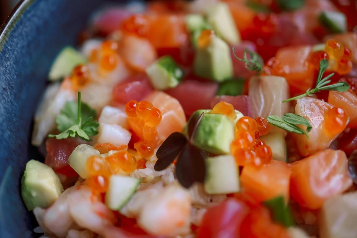 This vibrant bowl features diced raw fish, such as salmon and tuna, mixed with cubes of creamy avocado and resting on a bed of rice. The dish is accented with glistening orange salmon roe and delicate microgreens for a fresh, colorful presentation.