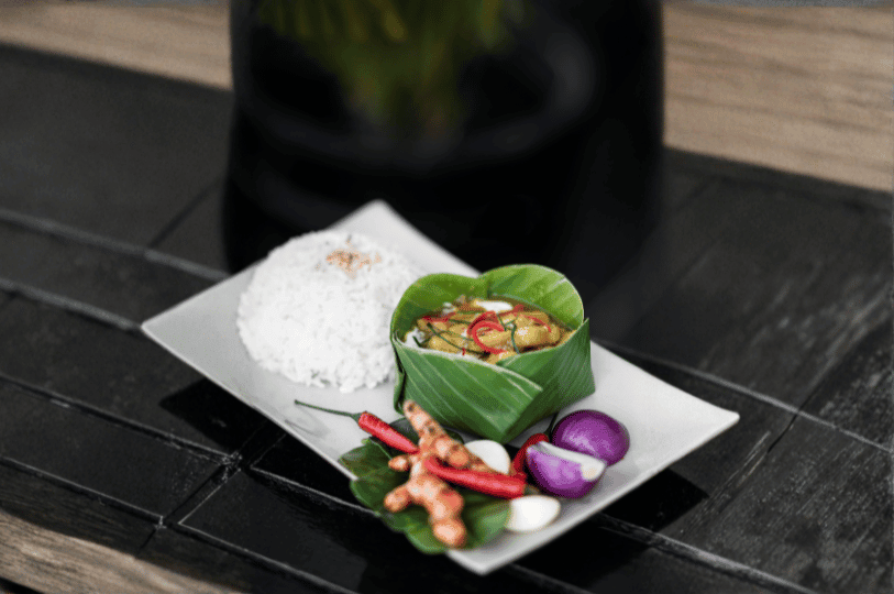 Cambodian fish amok in a banana leaf boat, topped with coconut cream and red chili, served alongside a small bowl of dark chili condiment on a rustic woven mat.