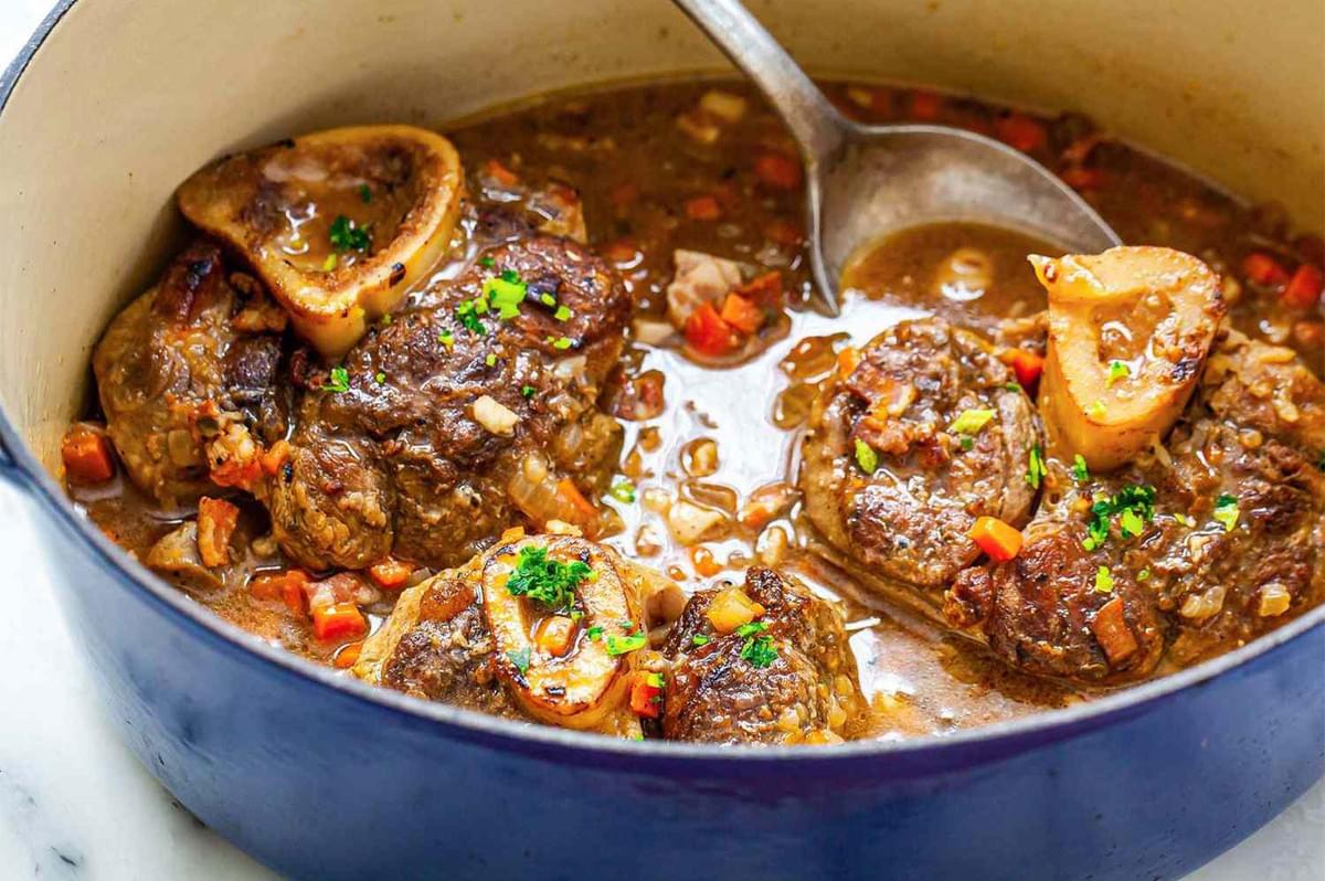 Close-up of Osso Buco (braised veal shanks) simmering in a rich, dark brown broth with diced vegetables (mirepoix) inside a blue Dutch oven, ready to be served.
