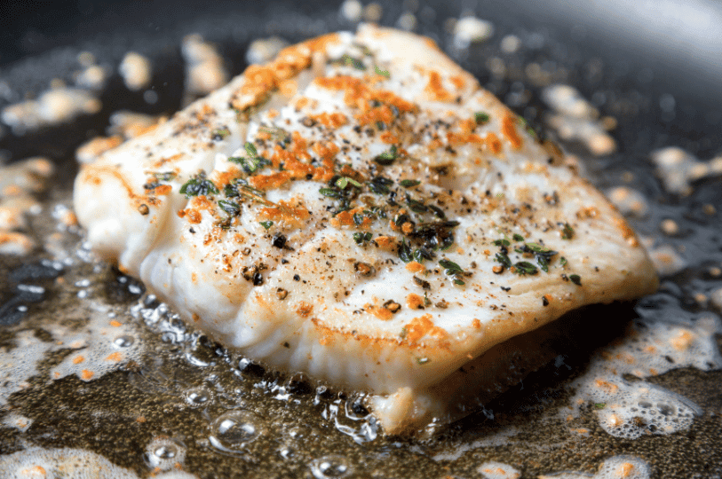 A close-up of a white fish fillet being pan-seared with fresh herbs and spices, featuring a delicate, crispy golden crust.
