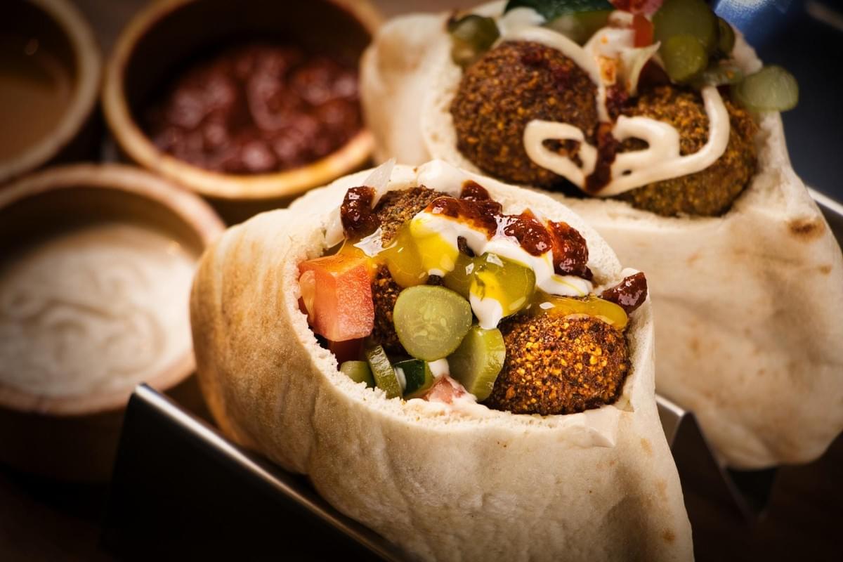 Two warm pita pockets filled with golden-brown falafel, diced tomatoes, sliced pickles, and drizzled sauces rest upright in a metal stand. Small wooden bowls filled with complementary sauces sit in the softly blurred background.