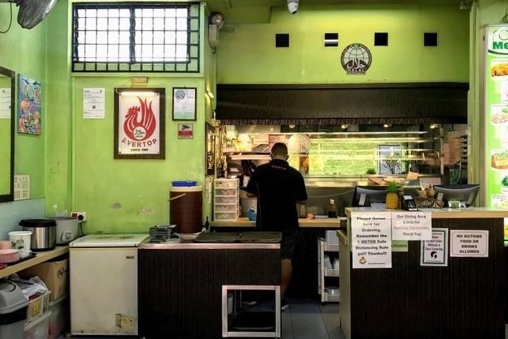  This image shows the interior of an Evertop Family Restaurant, which prominently displays its Halal Singapore certification on the lime green walls and above the counter. A staff member in a black uniform is seen working behind the service desk, which is marked with several notices regarding queuing, dining policies, and safety rules.
