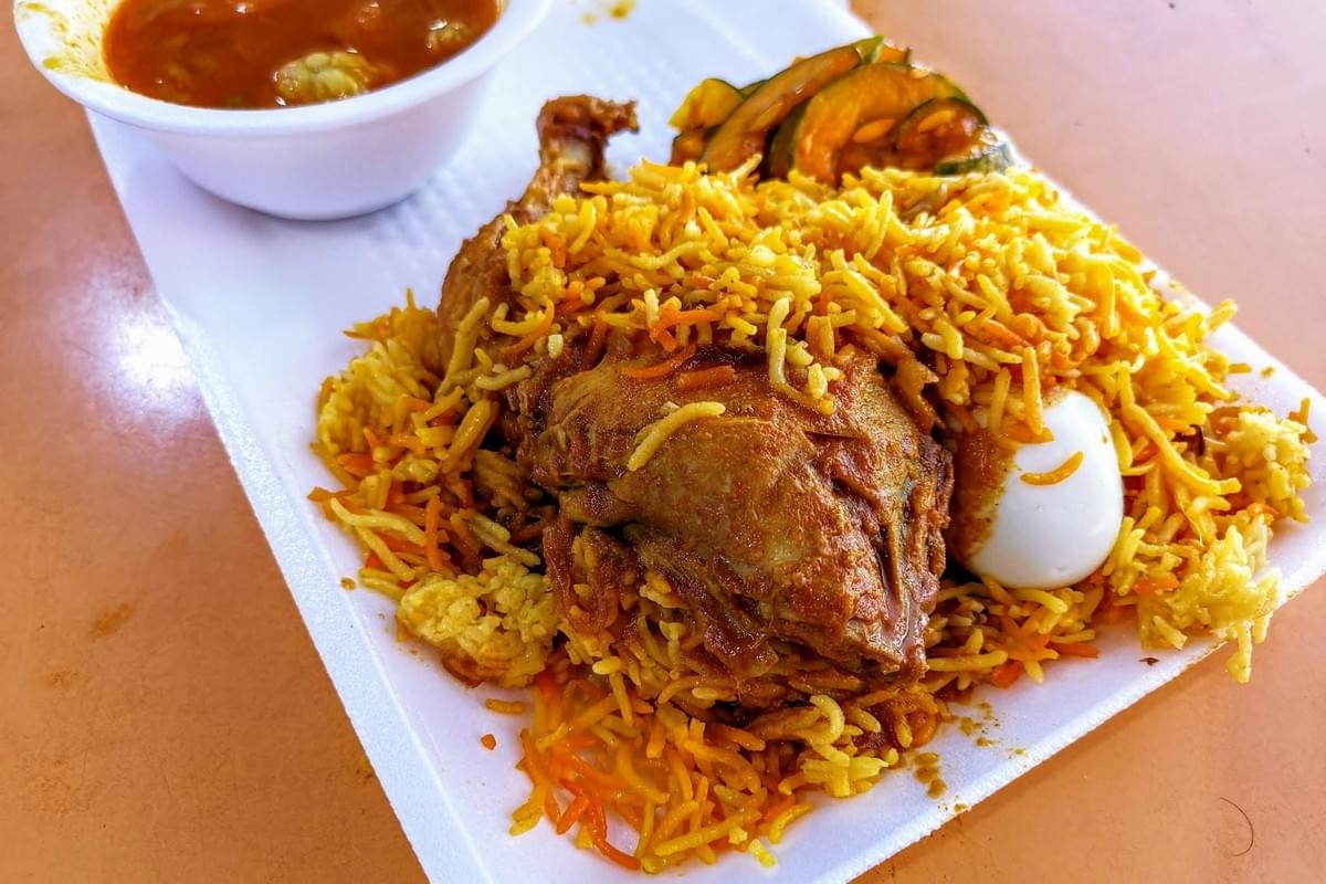 A generous portion of spiced chicken biryani is served on a white tray with a hard-boiled egg and a side of pickled cucumbers. A small white bowl of savory orange curry sits in the background to accompany the meal.
