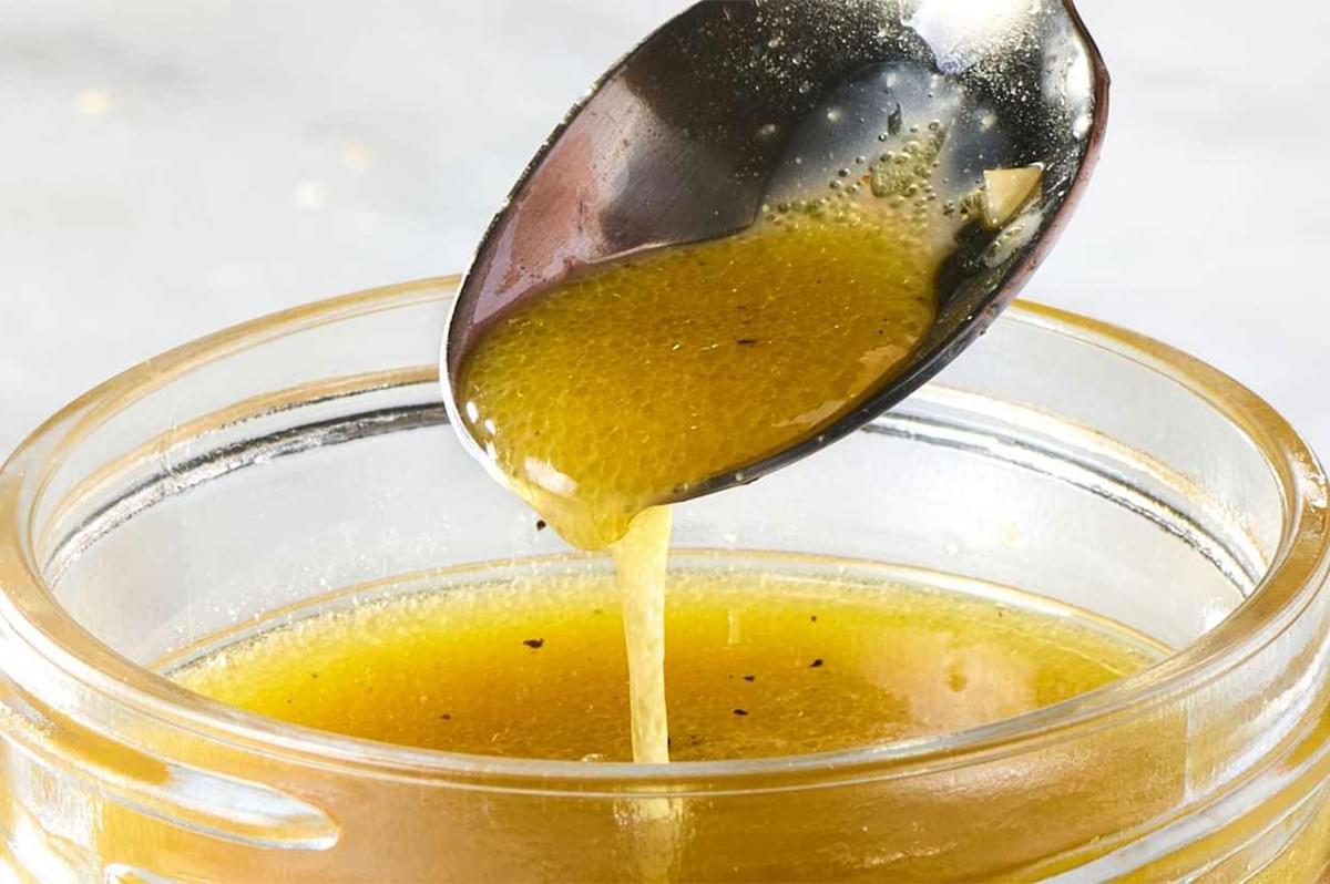 A spoonful of vibrant yellow vinaigrette dressing, speckled with herbs and pepper, dripping back into a clear glass jar on a white background.