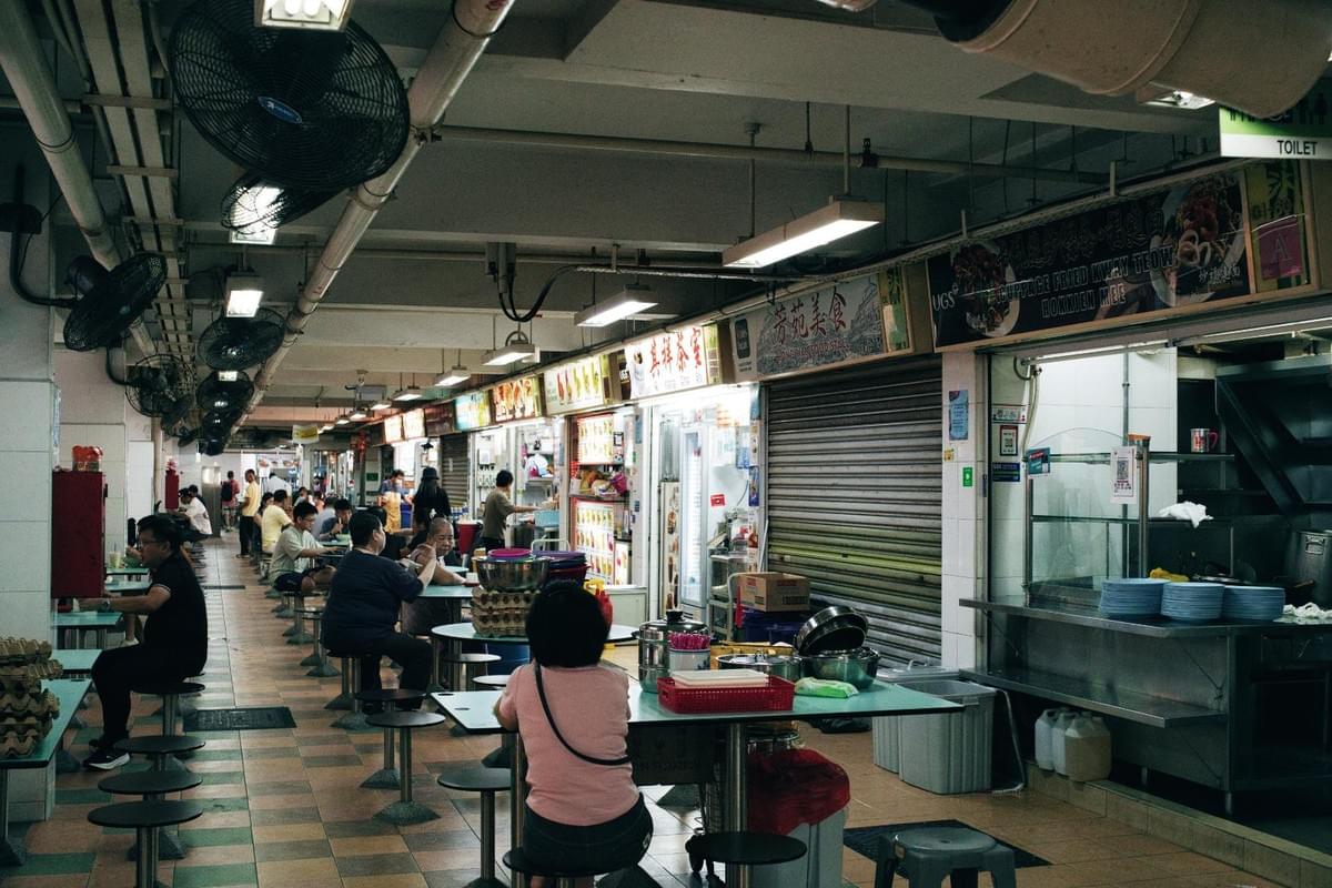 A bustling indoor Asian hawker center features a row of food stalls with bright Chinese signage, overhead fans, and exposed ceiling pipes. Diners sit at round metal tables on small stools eating and chatting, while a woman in a pink shirt sits in the foreground with her back to the camera.