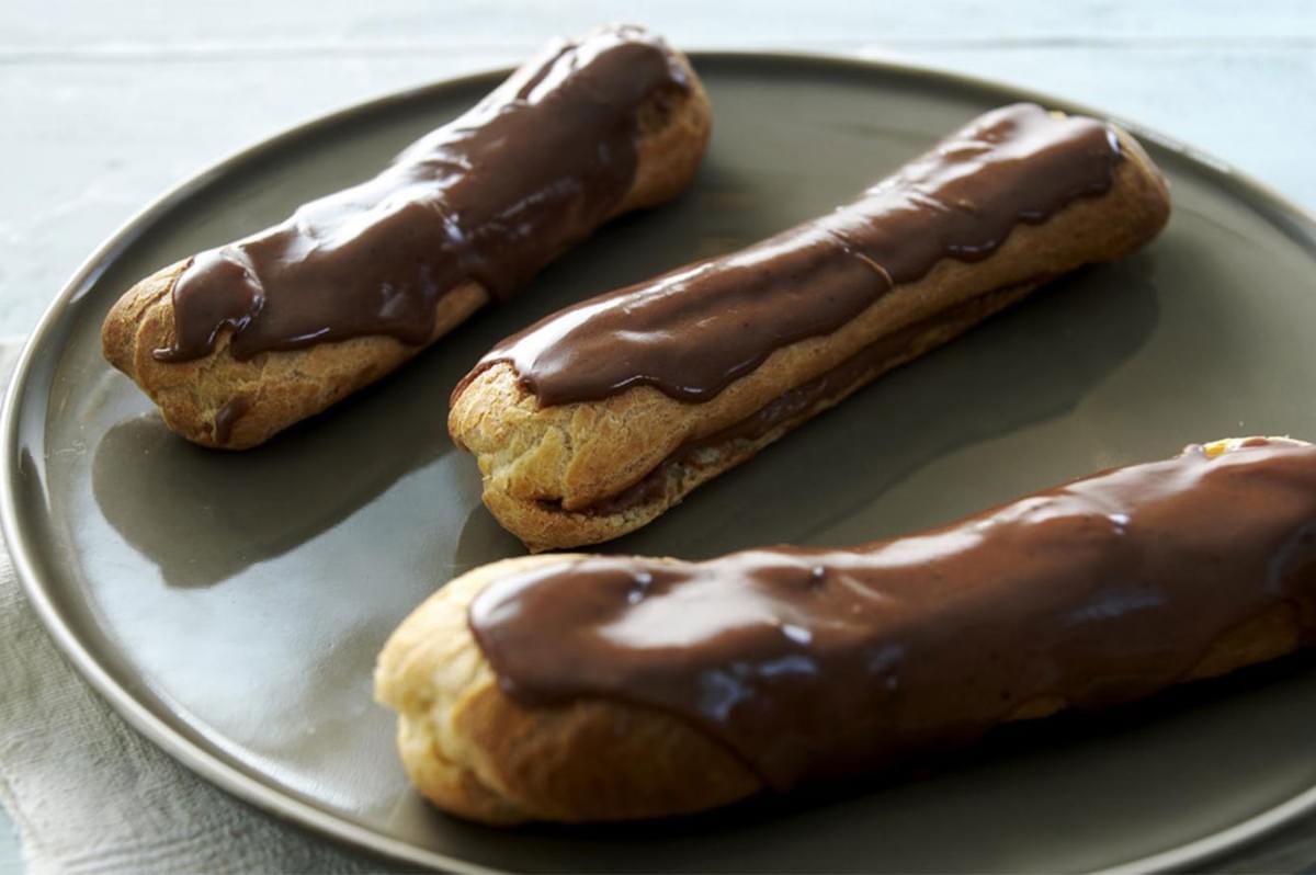 Three long, dark-chocolate glazed eclairs filled with pastry cream, resting on a round, matte grey plate against a light blue background.