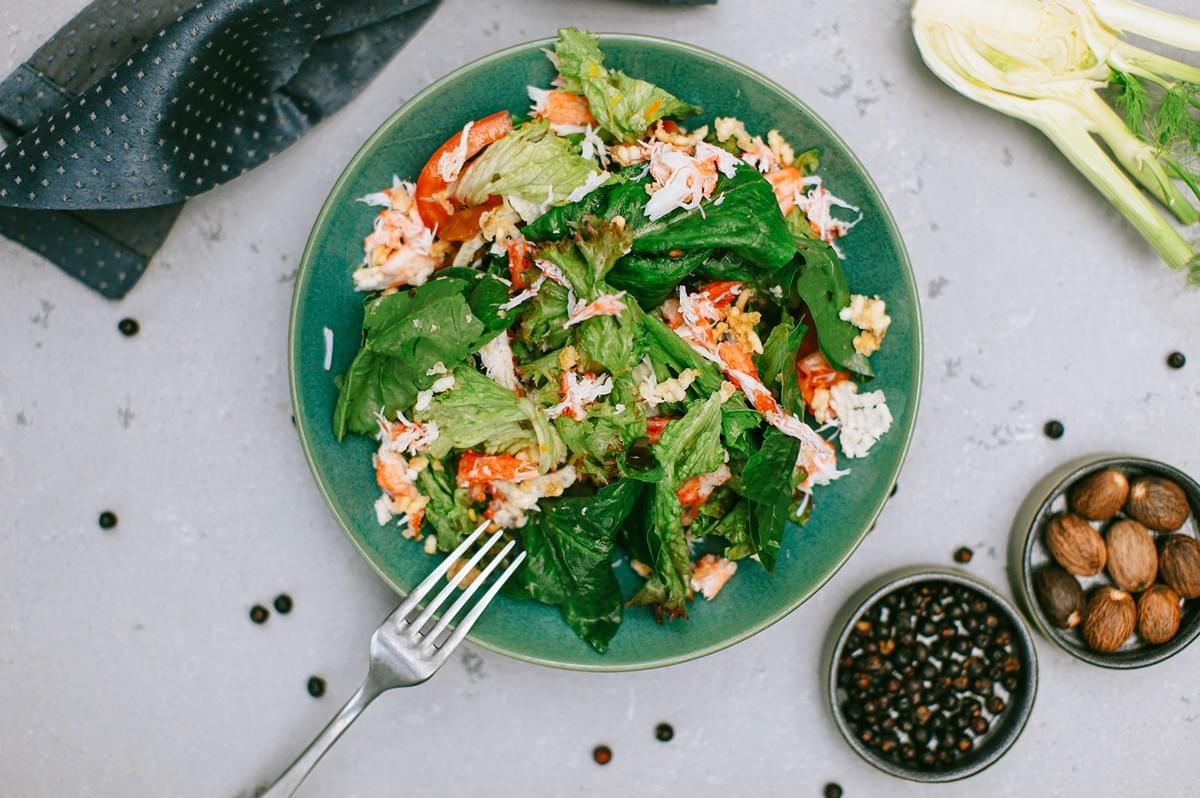 Fresh crab meat and spinach salad on a green plate next to a fork, fennel, and peppercorns.
