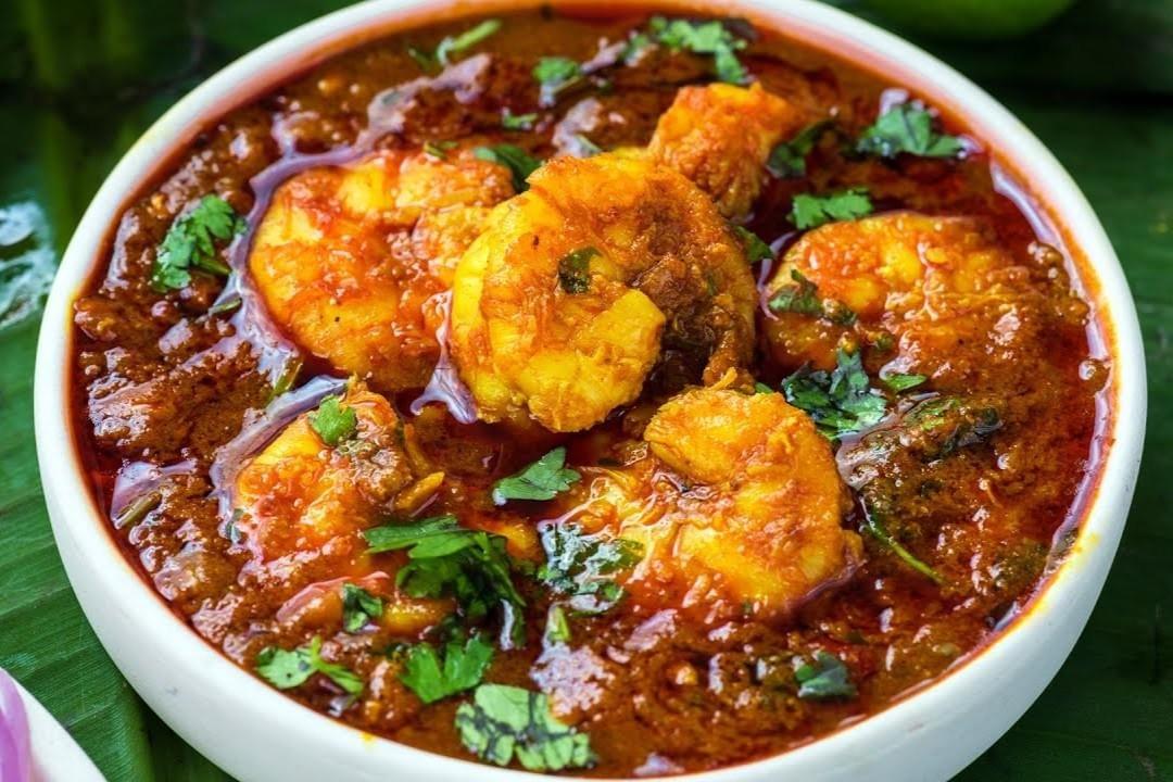 The image shows a white bowl filled with a rich, vibrant orange-red shrimp curry. The dish is garnished with fresh chopped cilantro and sits on a green banana leaf background.